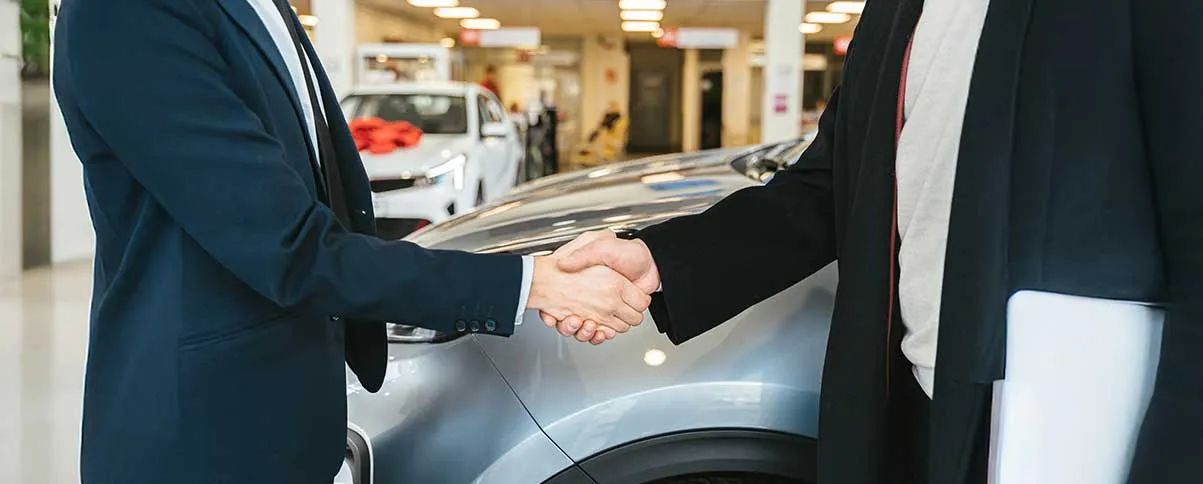 Cierre de trato con apretón de manos entre vendedor y cliente frente a un automóvil