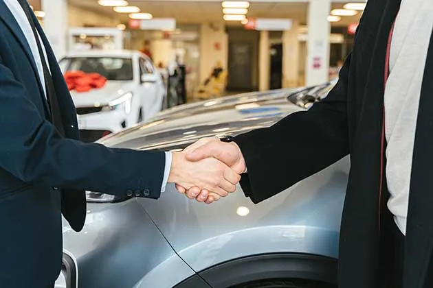 Cierre de trato con apretón de manos entre vendedor y cliente frente a un automóvil