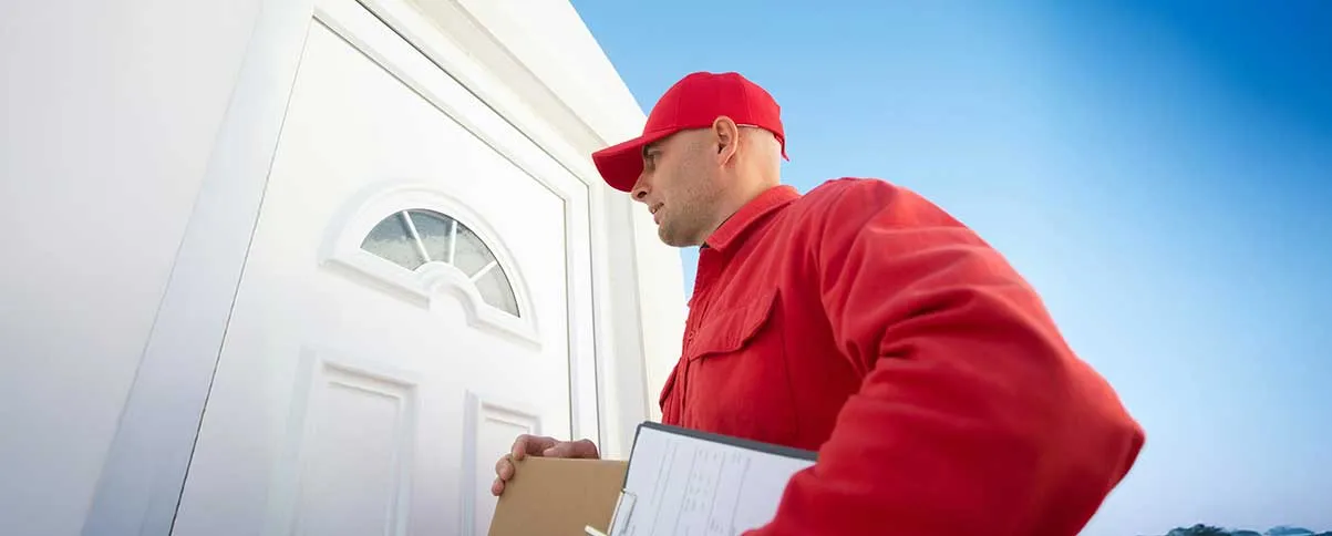 Repartidor con uniforme rojo tocando la puerta de una casa mientras sostiene un paquete y una libreta