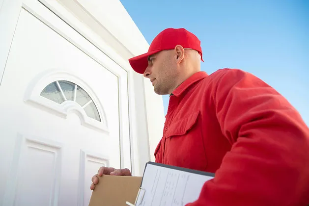 Repartidor con uniforme rojo tocando la puerta de una casa mientras sostiene un paquete y una libreta